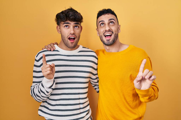 Junge hispanische schwule Paar steht über gelbem Hintergrund erstaunt und überrascht Blick nach oben und zeigt mit Fingern und erhobenen Armen.  - Foto, Bild