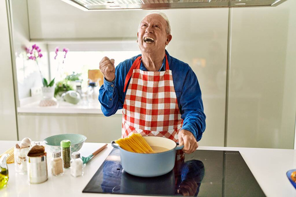 Senior άνθρωπος με γκρίζα μαλλιά μαγείρεμα σπαγγέτι στο σπίτι κουζίνα ουρλιάζοντας υπερήφανος, γιορτάζει τη νίκη και την επιτυχία πολύ ενθουσιασμένος με σήκωσε τα χέρια  - Φωτογραφία, εικόνα