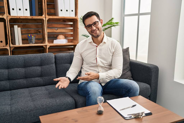 Young hispanic man with beard working at consultation office inviting to enter smiling natural with open hand  - Foto, Bild