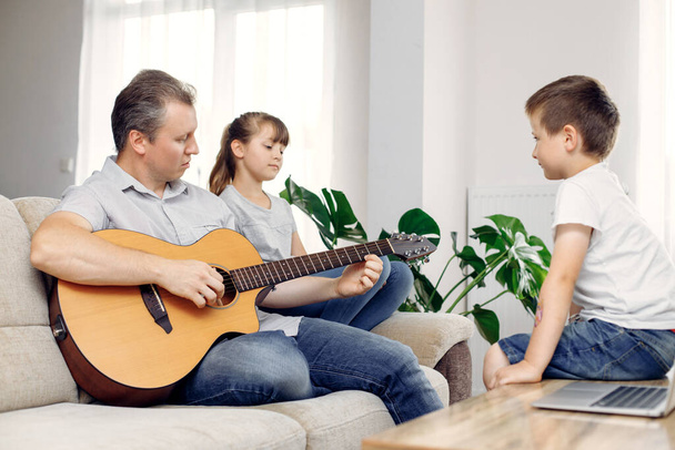 Tata i córka siedzą na kanapie, a tata gra na gitarze. Syn siedzi obok niego i słucha. Czas z rodziną. Wszyscy są szczęśliwi, że spędzają razem czas. - Zdjęcie, obraz