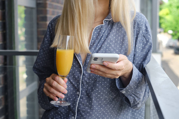 The girl holds a smartphone and drinks orange juice on the balcony. Fresh organic juice in a glass. Woman watching social networks, sending text messages or using apps on her mobile phone - Foto, Bild