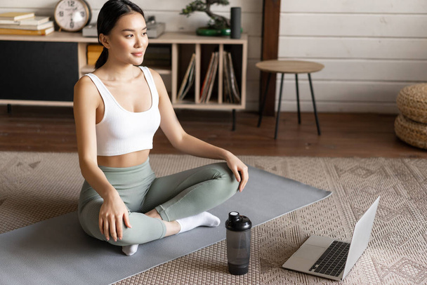 Sport and lifestyle concept. Smiling young asian woman doing yoga at home, follow meditation course on laptop, sitting in zen pose on floor mat. - Photo, Image