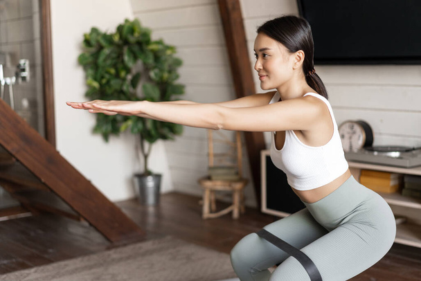 Schattig aziatisch fitness meisje thuis doen workout, hurken met stretching elastische touw op haar benen, oefenen voor fit en gezond lichaam. - Foto, afbeelding