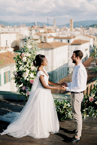 Bride and groom are standing near the wedding semi-arch of flowers on the terrace of the building. High quality photo - Photo, image