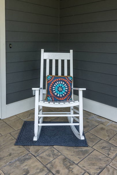 Μια σφραγισμένη τσιμεντένια μπροστινή βεράντα με λευκή κουνιστή καρέκλα με πολύχρωμο μαξιλάρι. - Φωτογραφία, εικόνα