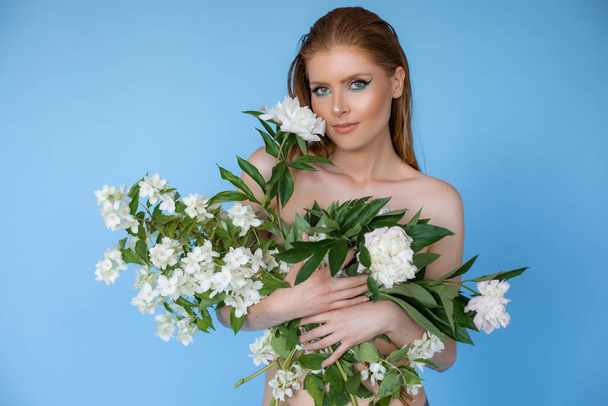 Close up portrait of confident, serious young blonde naked woman looking at camera with white flowers in hands, covering her body on blue background. Health and bode care, perfect skin spa treatment - Fotoğraf, Görsel