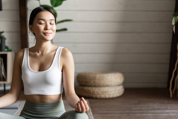Mindfulness en meditatie concept. Jonge aziatische vrouw doet workout thuis, yoga meditatie in de woonkamer op vloermat, steeds gefocust. - Foto, afbeelding