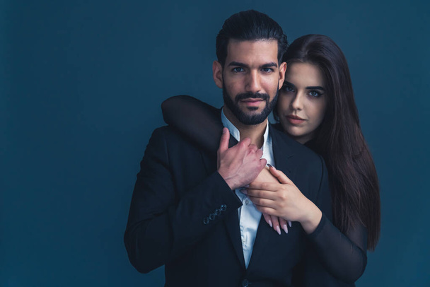 Hispanic man with beard wearing suit hugged by girlfriend from the back putting his hand on her arms looking seriously into camera. Dark background studio shot. High quality photo - Foto, immagini