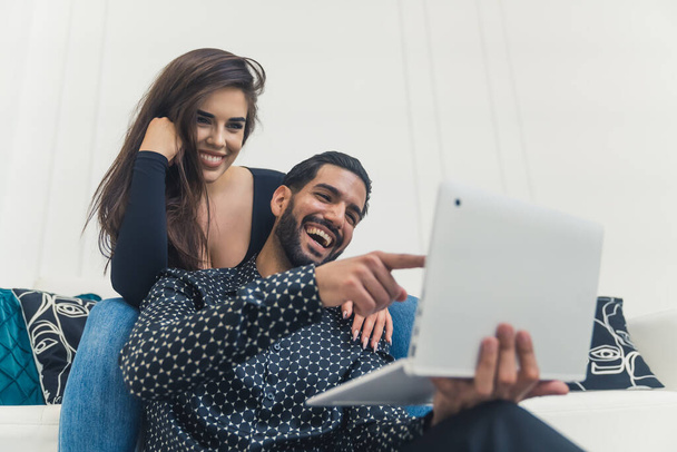 Dark-haired man with beard laughing and pointing at laptop with his girlfriend sitting on couch smiling and leaning on him. Indoor shot. High quality photo - Photo, image