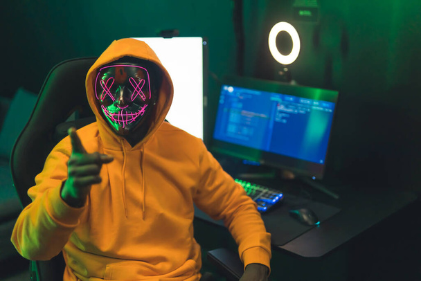 Young black man in orange hoodie and strange mask pointing at camera sitting by desk with gaming setup and ring light. Dark green lighting. High quality photo - Foto, immagini