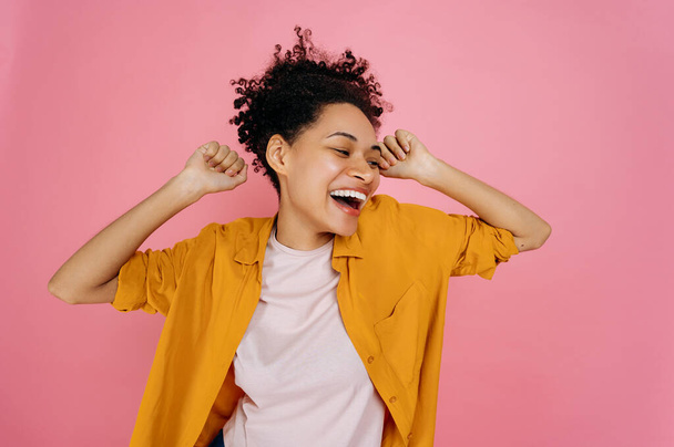 Happy cheerful african american girl in casual wear, dancing to favorite music, having fun, rejoice in luck, celebrating win, smiling, stand on isolated pink background. Joy, victory, luck, dancing - Foto, Imagen