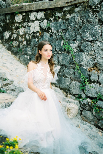 Bride in a white dress sits on the stone steps near the wall of the building. High quality photo - Foto, Bild