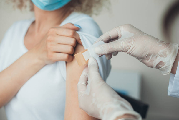 Lääkäri suojakäsineissä laittaa liiman nuorten naisten käsivarteen rokotuksen jälkeen. Rokotus sepelvaltimovirusta vastaan COVID-19-pandemian aikana - Valokuva, kuva