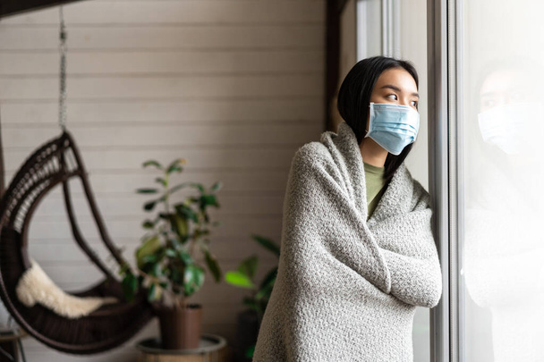 アジアの女の子は病気になる,顔の医療マスクを身に着けています, covid-19隔離に滞在し、外を見ます,徒歩で行くことへのあこがれ. - 写真・画像