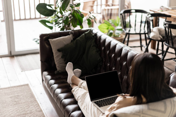Visão traseira da mulher asiática sentada em casa e descansando na sala de estar, deitada no sofá com laptop, assistindo vídeos online. - Foto, Imagem