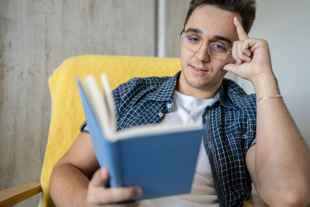 Один чоловік кавказький чоловік молодий дорослий сидить у кріслі вдома, читаючи книгу, носить блакитну сорочку і носить окуляри, думаючи про споглядання дозвілля концепція навчання копіювати простір
 - Фото, зображення