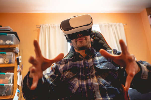 Handsome and happy young latino man with plaid shirt playing with virtual reality helmet (VR glasses) on a gamer chair with luminous window behind in orange bedroom on home - Foto, Imagen