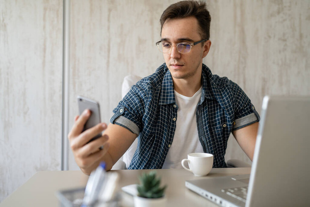 One man caucasian male sitting in chair in the office at work at desk using mobile phone to make a call while working on project wear blue shirt and eyeglasses talking consulting copy space - Foto, imagen