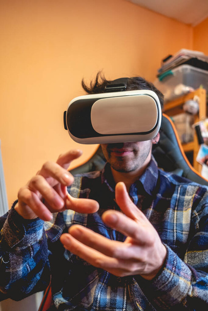 Handsome and happy young latino man with plaid shirt playing with virtual reality helmet (VR glasses) on a gamer chair in orange bedroom on home - Photo, Image