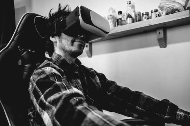 Handsome and happy young latino man with plaid shirt playing with virtual reality helmet (VR glasses) on a gamer chair in orange bedroom on home (in black and white) - Foto, Imagen