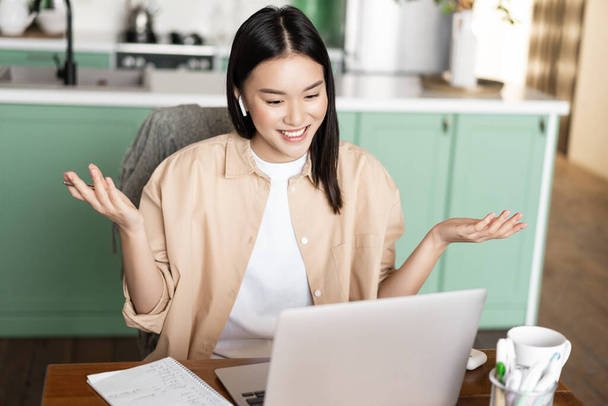 Aziatische vrouw tutor met online lessen van thuis, werken via laptop, het hebben van video call conferentie, het dragen van een koptelefoon, zitten aan tafel omringd door studiemateriaal. - Foto, afbeelding