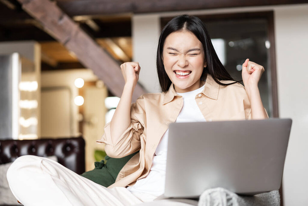Mulher asiática comemorando, aplaudindo de boas notícias no laptop, triunfando da alegria, sentada na sala de estar em casa. - Foto, Imagem