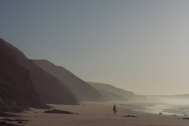 A silhouette of a woman walking along the beach surrounded by huge rock formation on a misty day - Foto, afbeelding
