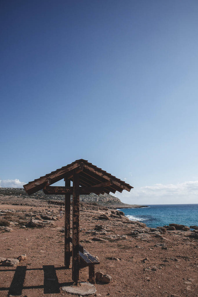 Όμορφη παραλία με ξύλινο παγκάκι με στέγη σε πρώτο πλάνο του βουνού Cape Greco στην Αγία Νάπα, Κύπρος.  - Φωτογραφία, εικόνα