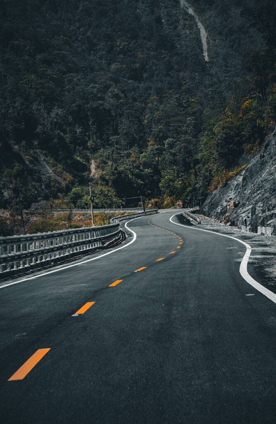 A vertical shot of an empty curvy road at the bottom of a forested hill - Foto, imagen