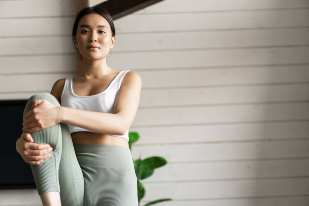 Portret van Aziatisch meisje dat thuis traint, benen optilt en afstand bekijkt, in de woonkamer traint zonder fitnesstoestel. - Foto, afbeelding
