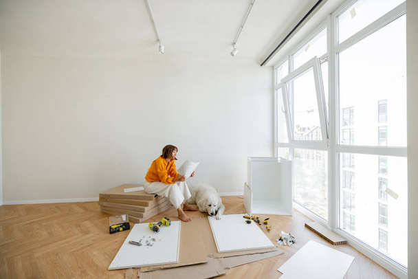 Young woman assembles furniture by herself, sitting with dog near window and reading manual at new apartment. Wide view with copy space - Photo, Image