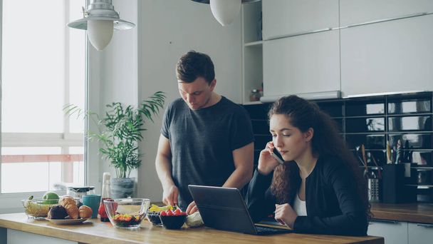 serious caucasian nice couple in kitchen, man is cutting salad, he is standing silent, woman is sitting at table working on laptop computer and talking on phone - Photo, image