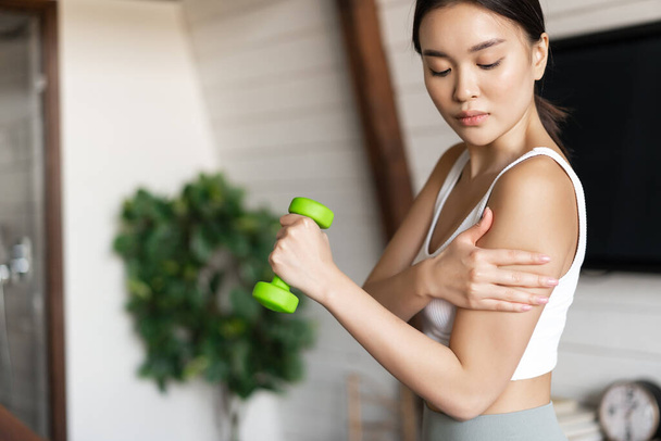 Aktivní a zdravé asijské dívka s fit tělo dělá fitness cvičení doma, zvedání činky a dotýkání se biceps, cvičení v obývacím pokoji. - Fotografie, Obrázek