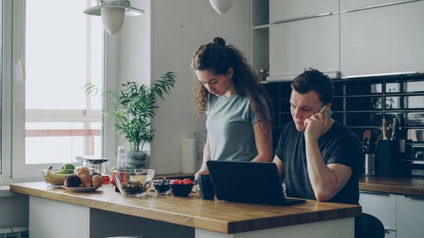 serious couple in kitchen, woman is cutting salad, she is standing silent, man is sitting at table working on laptop and talking on phone, he is annoyed and angry - Zdjęcie, obraz