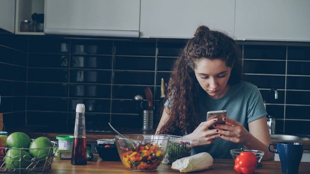 Retrato de una joven hermosa mujer caucásica sentada a la mesa en la moderna cocina espaciosa y luminosa, usando un teléfono inteligente, mensajes de texto, platos con comida están delante de ella
 - Foto, imagen