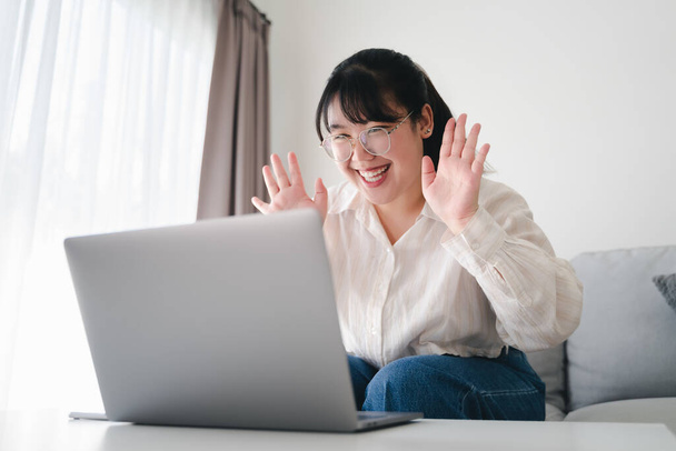 Young Asian woman using laptop computer for online video conference call waving hand making hello gesture on the couch in living room. - Fotografie, Obrázek