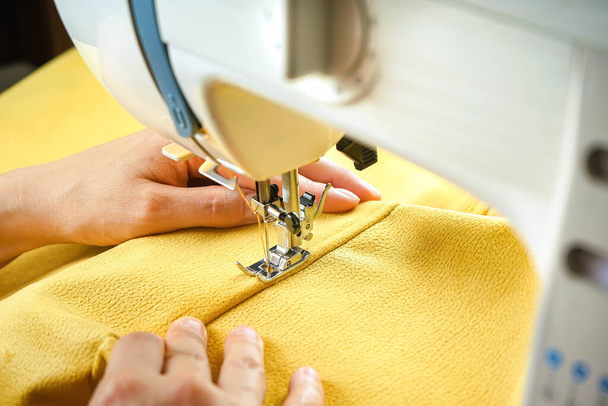 Terzi kadın el ele tutuşuyor ve işyerindeki modern dikiş makinesine sarı tekstil kumaşı dikiyor. Dikiş, döşeme, giysi, onarım, DIY. El yapımı, hobi, küçük işletme konsepti - Fotoğraf, Görsel