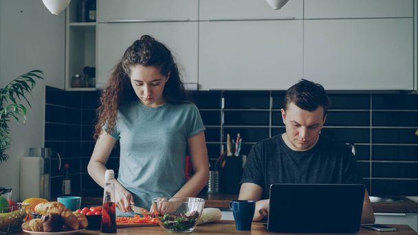 young couple at table, man is sitting working on laptop, he is concentrated and pensive, beautiful curly woman is standing cooking cutting vegetables for salad, she is seriuos - Photo, image