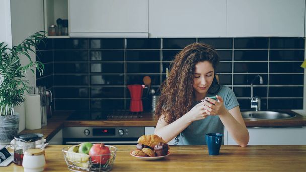 young beautiful curly pretty caucasian woman sitting at table in nice modern kitchen using smartphone, she is texting someone and smiling, calm, happy and positive - Fotografie, Obrázek