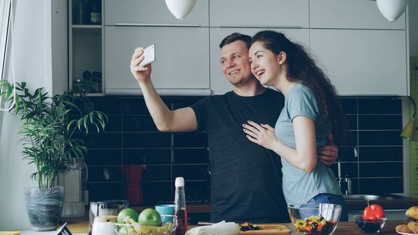 young happy couple having online video call with smartphone camera while cooking in the kitchen at home - Φωτογραφία, εικόνα