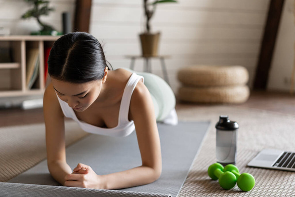 Sportliches asiatisches Mädchen in Aktivkleidung beim Workout zu Hause, Videosportführer, Online-Fitnesstrainer am Laptop, in Planke auf Bodenmatte stehend. - Foto, Bild