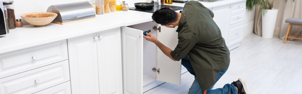 Young muslim man with screwdriver fixing cabinet under worktop in kitchen, banner  - Foto, immagini