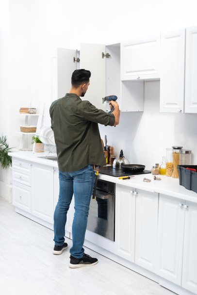 Muslim man using electric screwdriver while fixing kitchen cupboard  - Zdjęcie, obraz