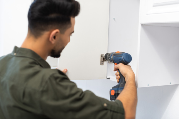 Blurred arabian man with electric screwdriver fixing hinge in kitchen cupboard  - Foto, imagen
