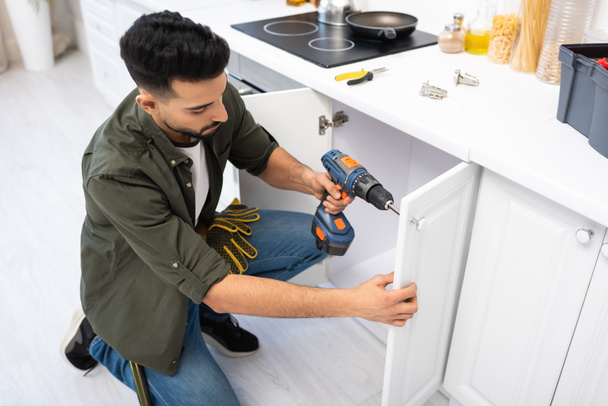 Overhead view of arabian man with electric screwdriver fixing door of cabinet near tools in kitchen  - Foto, afbeelding