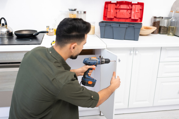 Young arabian man with electric screwdriver fixing door of kitchen cabinet near toolbox on worktop  - Fotó, kép
