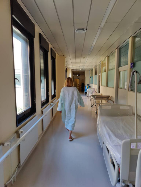 Female patient wearing hospital robe walking in long empty hospital hallway - Zdjęcie, obraz