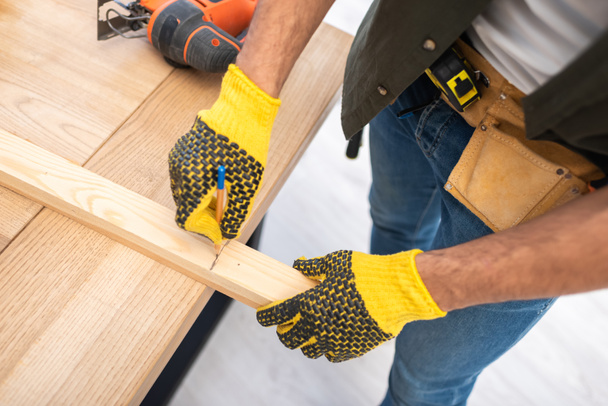 Cropped view of man in gloves and tool belt marking wooden plank at home  - Foto, imagen