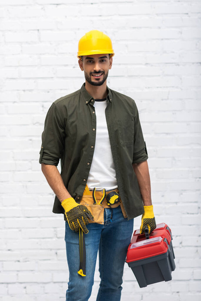 Positive muslim man in hardhat holding toolbox at home  - Foto, immagini