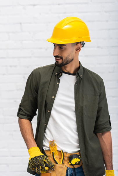 Smiling muslim man in hardhat and gloves touching tool belt at home  - Zdjęcie, obraz